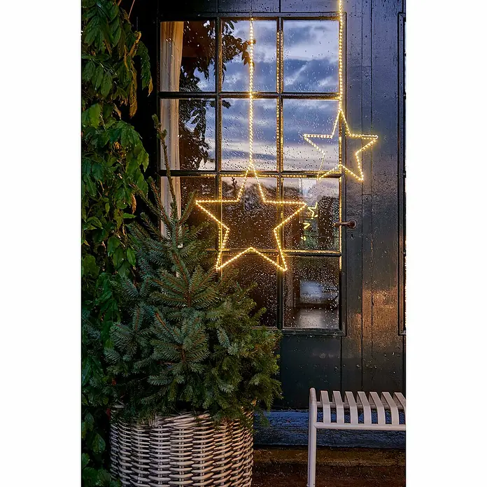 Guirnalda luminosa LED estrella - Para exterior, Diámetro: 42 cm, Color de luz: Blanco cálido, Número de bombillas: 130 ud. - Vista exterior con decoraciones de estrellas iluminadas, un abeto en una cesta y un banco. Las estrellas emiten un brillo cálido frente a una puerta lluviosa.