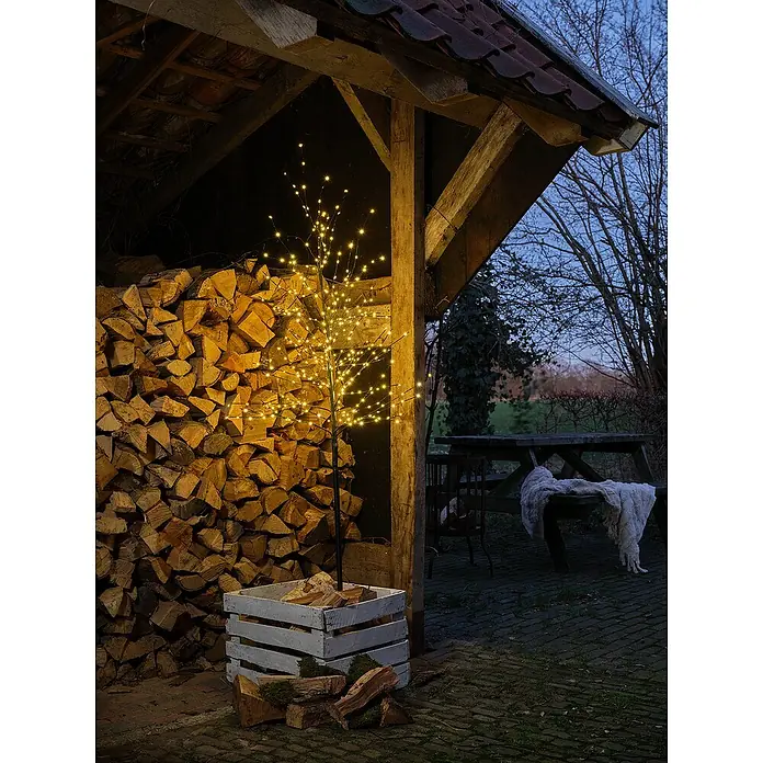 Árbol de Navidad de LEDs - Para interior, 480 ud., Color de luz: Blanco cálido - Escena decorativa de árbol de luz exterior con pila de leña y zona de estar al fondo, creando un ambiente cálido y acogedor.