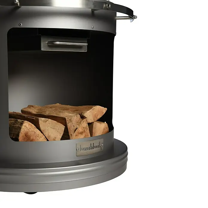 Buschbeck Grillkamin Auckland - Hauptgrillfläche: Ø 42,5 cm, Edelstahl, Dunkelgrau - Nahaufnahme eines modernen Stahlgrills mit Holzscheiten im Lagerbereich, der ein elegantes Design und Funktionalität vereint.
