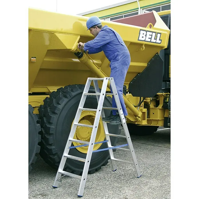 Krause Stabilo Doppelleiter - Anzahl Tritte: 14 Stk., Arbeitshöhe: 3,2 m, Aluminium - Arbeiter mit blauer Arbeitskleidung und Schutzhelm auf einer Aluminium-Doppelstufenleiter vor einem gelben Bell Baufahrzeug.