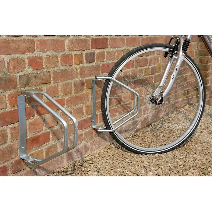 Mottez Aparcamiento de bicicletas - L x An x Al: 285 x 85 x 328 cm, Acero, Montaje en la pared - Portabicicletas de pared. El portabicicletas plateado está montado en una pared de ladrillo y sujeta una bicicleta.