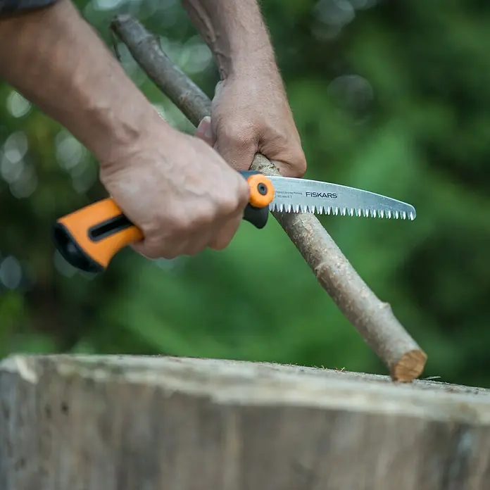 Fiskars Hand-Astsäge Xtract SW73 - Länge: 22,5 cm, Max. Aststärke: 12 mm, Stahl, Orange/Schwarz - Nahaufnahme einer Fiskars-Handsäge beim Schneiden eines Astes. Das Werkzeug liegt auf einem Baumstumpf und der Hintergrund ist verschwommen.