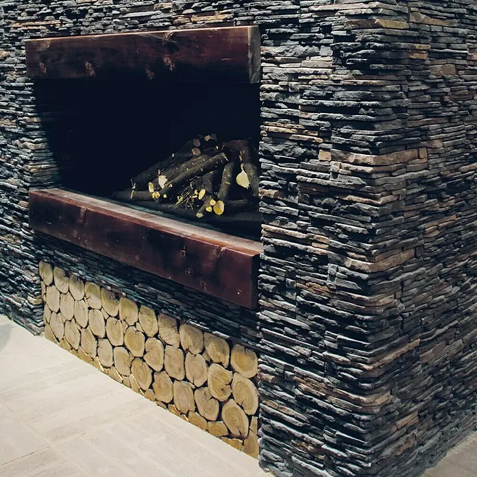 Esquina exterior Galicia PIzarra - 20 x 12,5 x 10 cm, Antracita - Primer plano de una chimenea hecha de piedra y troncos de madera. La chimenea oscura es rústica y ofrece un ambiente acogedor.