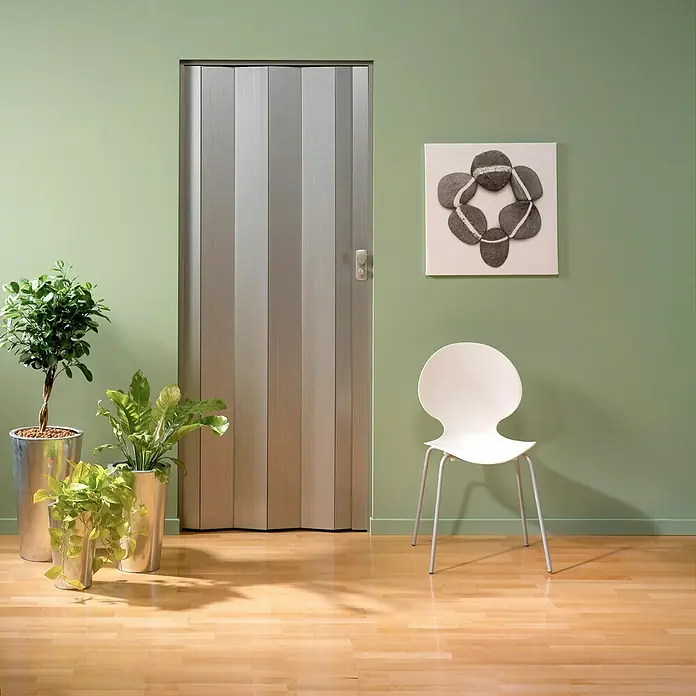 Grosfillex Puerta plegable Spacy - 84 x 205 cm, Plata - La imagen muestra una puerta plegable plateada que se integra perfectamente en el interior moderno, con plantas y una silla blanca.