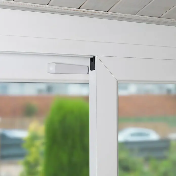 Inofix Tope de cierre 5201 - Blanco, Plástico, Apto para: Puertas y ventanas correderas - Primer plano de un marco de ventana blanco con un mecanismo de bloqueo negro. La ventana ofrece una vista de un paisaje exterior borroso.