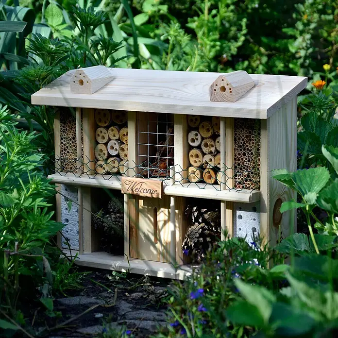 Dobar Insektenhotel Landsitz Superior - 47 x 12,5 x 34 cm, Holz, Natur, Kiefer - Ein hölzernes Insektenhotel mit verschiedenen Füllungen, umgeben von grüner Vegetation, bietet einen Unterschlupf für nützliche Insekten im Garten.