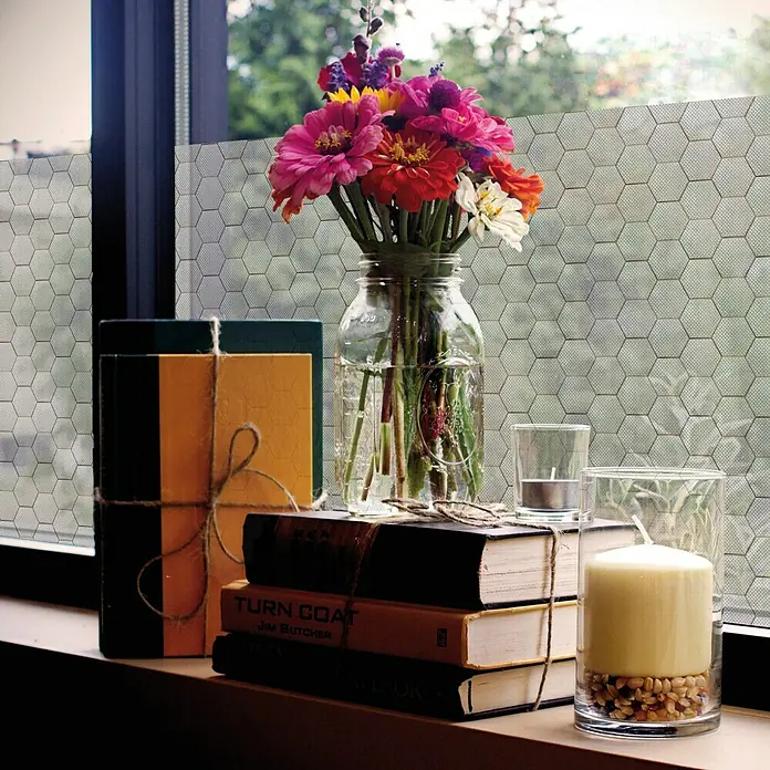 Lámina para ventana gofrado Fix hexagon mini - 92 cm x 1,5 m, Transparente - Naturaleza muerta con flores en un jarrón tipo mason jar, libros, velas y cajas decorativas frente a una ventana con un patrón hexagonal.