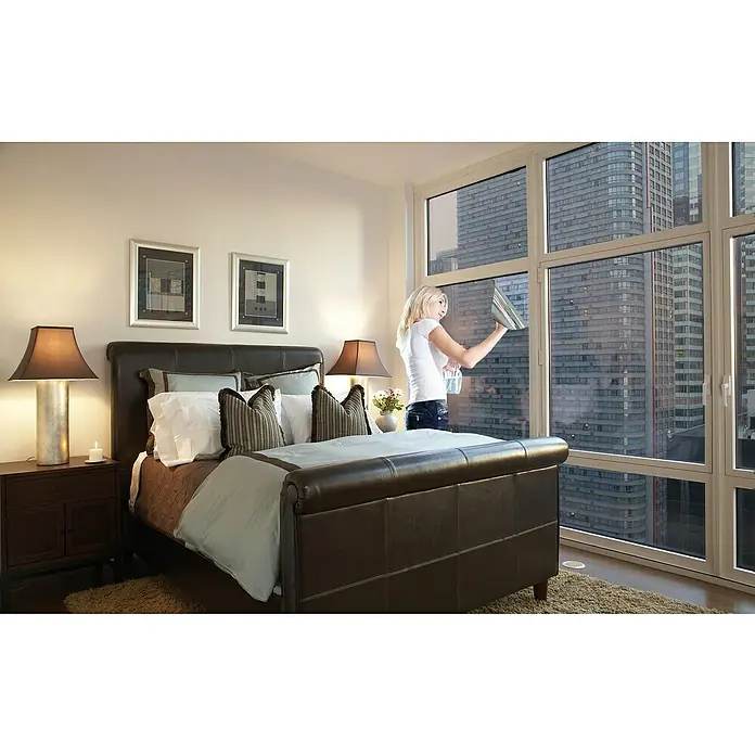 Lámina para ventana estático Efecto Espejo - 75 x 150 cm, Efecto espejo, Plateado - Un dormitorio con una cama grande y una mujer limpiando las ventanas para disfrutar de la vista de la ciudad.