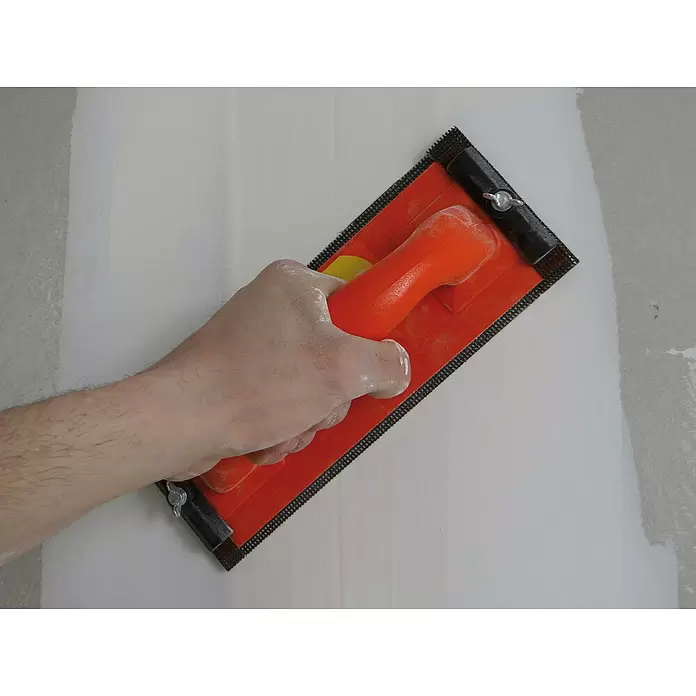 Mondelin Handschleifer - 240 x 80 mm, Geeignet für: Gipsplatten - Nahaufnahme einer Hand, die einen orangefarbenen Trockenbau-Handschleifer mit schwarzem Schleifgitter an einer weißen Wand verwendet.