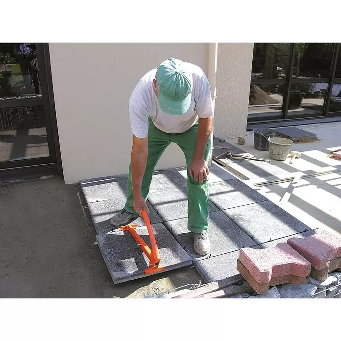 Mondelin Plattenheber - Stahl - Oranges Plattenheber-Werkzeug, von einem Arbeiter zum präzisen Verlegen grauer, gesprenkelter Terrassenplatten verwendet.