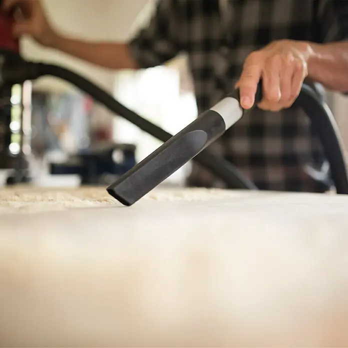 Herkules Elektro-Nass- & Trockensauger H-NT 50 Inox - 1.600 W, 50 l - Ein Mann benutzt einen Staubsauger mit Aufsatz, um einen Holztisch von Sägespänen zu reinigen. Die Werkstatt wirkt aufgeräumt.