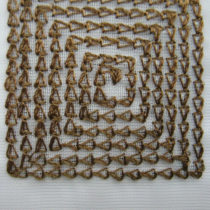 Elbersdrucke Bistrogardine Quadro - 140 x 48 cm, 100 % Polyester, Braun/Weiß - Nahaufnahme einer detaillierten Kreuzsticharbeit mit einem quadratischen Spiralmuster in einem warmen Braunton auf hellem Stoff.