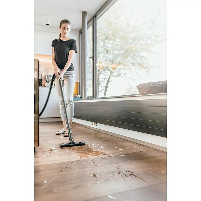 Kärcher Aspirador en seco y húmedo eléctrico WD 2 Plus - 1.000 W, Volumen de recipiente: 12 l, Con bolsa - Una mujer usa una aspiradora Kärcher para limpiar un piso de madera donde se ha derramado líquido y fragmentos de vidrio.