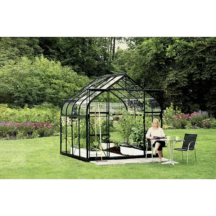 Vitavia Gewächshaus Dione 6700 - Außenmaß inkl. Dachüberstand (B x T): 263,5 x 258 cm, 3 mm, Schwarz - Geräumiges Gewächshaus mit Stahlrahmen und Glaswänden. Eine Frau sitzt am Tisch daneben und liest. Perfekt für Gartenliebhaber.