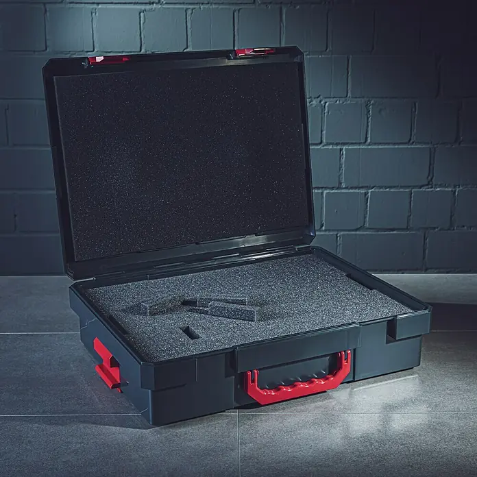 Wisent b-boXx Schaumstoffeinlage - Passend für: Wisent b-boXx - Bild eines offenen schwarzen Werkzeugkoffers mit rotem Griff, der mit Schaumstoff ausgekleidet ist, auf einer gefliesten Oberfläche vor einer Ziegelwand.