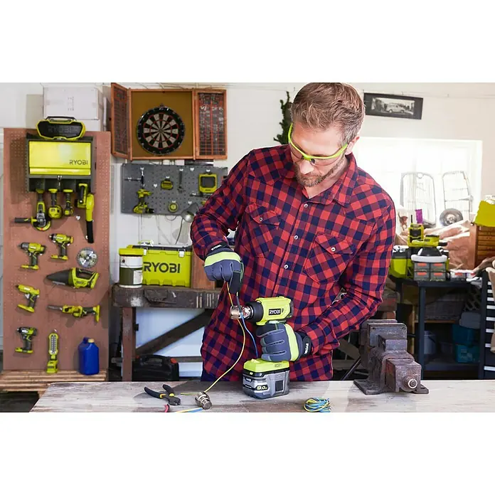 Ryobi ONE+ Akku-Heißluftgebläse R18HG-0 - 0 °C - 470 °C, Ohne Akku - Ein Mann benutzt eine Ryobi Akku-Heißluftpistole, um an Drähten zu arbeiten, mit Werkzeugen und Werkbänken im Hintergrund.