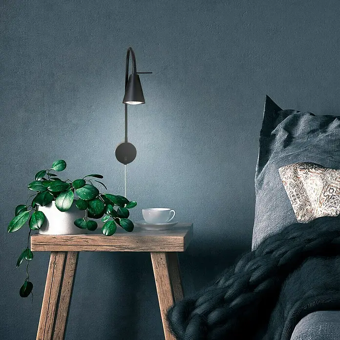 Briloner Foco de pared LED Comfort Light - Negro, 4 W - Una lámpara de pared negra moderna con brazo ajustable ilumina una planta de interior y una taza sobre una mesa de madera junto a una cama.