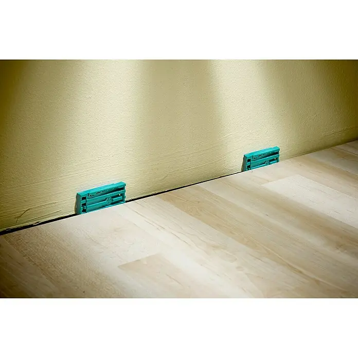 Wolfcraft Keile - 30 Stk. - Das Bild zeigt zwei türkisfarbene Abstandshalter zwischen einer beigen Wand und einem hellen Holzboden, die zur korrekten Installation dienen.