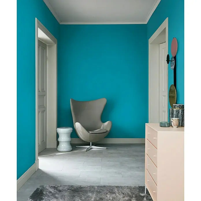 SCHÖNER WOHNEN-Farbe Wandfarbe Trendfarbe - Lagune, Matt, 2,5 l, Konservierungsmittelfrei - Einladender Raum mit türkisblauen Wänden, einem grauen Sessel und einer rosa Kommode, der eine moderne und stilvolle Atmosphäre schafft.