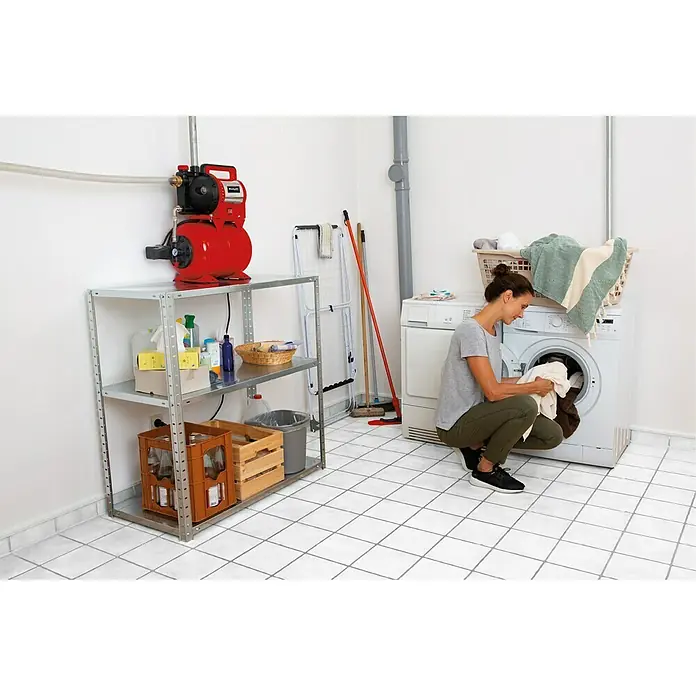 Einhell Hauswasserwerk GC-WW 1045 N - 1.050 W, Max. Fördermenge: 4.500 l/h, Max. Druck: 3 bar - Das Bild zeigt ein Hauswasserwerk auf einem Regal in einem Waschraum. Eine Frau hockt vor einer Waschmaschine und sortiert Wäsche.