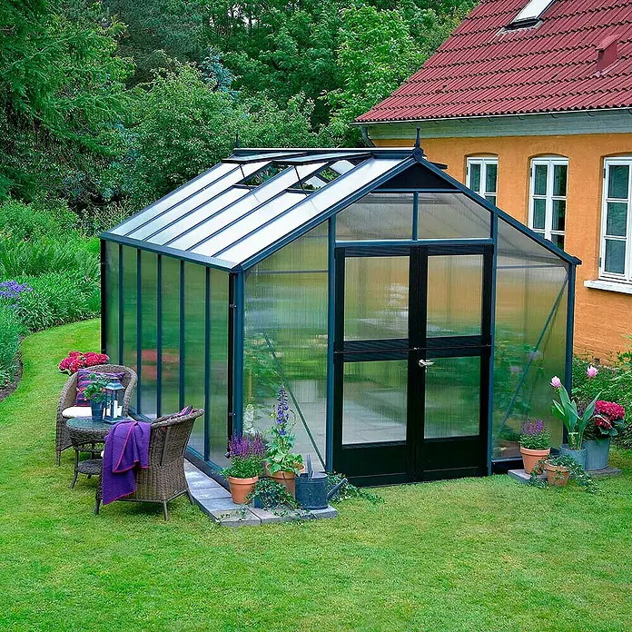 Juliana Gewächshaus Premium - Außenmaß inkl. Dachüberstand (B x T): 296 x 439 cm, Material Seitenwand: Kunststoff, 10 mm, Anthrazit/Schwarz - Ein elegantes Gewächshaus mit schwarzem Rahmen steht in einem gepflegten Garten neben einem Wohnhaus. Es ist hell und bietet viel Platz für Pflanzen.