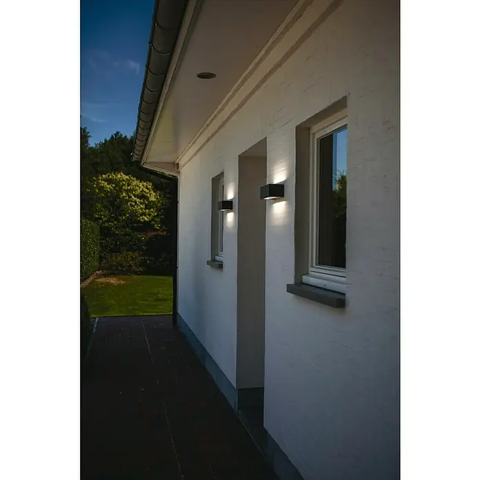 Lutec Aplique exterior LED Gemini - 42 W, 12 x 30 x 10,8 cm, Antracita, IP54 - Primer plano de una lámpara de pared exterior que emite un brillo cálido, montada en una pared de ladrillo blanco junto a una ventana con marco gris.