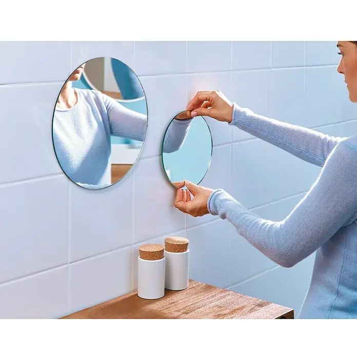 Tesa Powerbond Cinta de doble cara Resistente humedad - 1,9 cm x 1,5 m, Blanco - Una mujer fija un espejo redondo a una pared de azulejos sobre un estante de madera. Los espejos y los azulejos reflejan la luz.