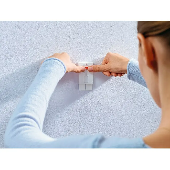 Tesa Clavo adhesivo ajustable - Blanco, Carga soportada: 2 kg, 2 ud. - Primer plano de una persona que fija un soporte de pared blanco con un deslizador rojo a una pared blanca, enfatizando su fácil instalación.