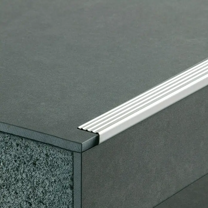 Escalón autoadhesivo - 1 m x 35 mm x 13 mm, Acero - Primer plano de un perfil de aluminio para escaleras sobre una escalera de piedra oscura, diseñado para proteger el borde y ofrecer resistencia al deslizamiento.
