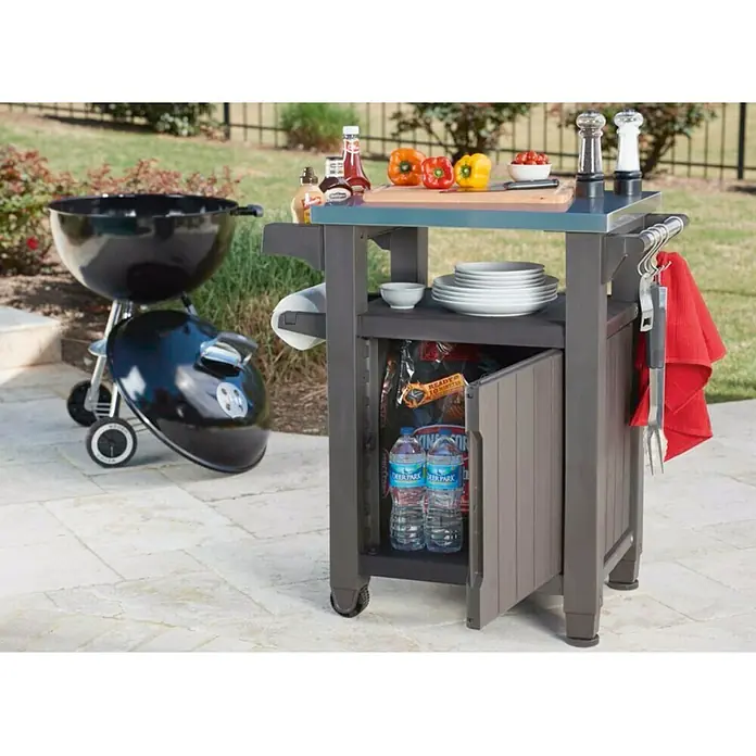 Keter Mesa auxiliar de jardín Unity - L x An: 82 x 52 cm, Marrón - Imagen de una estación de parrilla con parrilla de carbón, utensilios para parrilla, platos, salsas y botellas de agua en un patio.