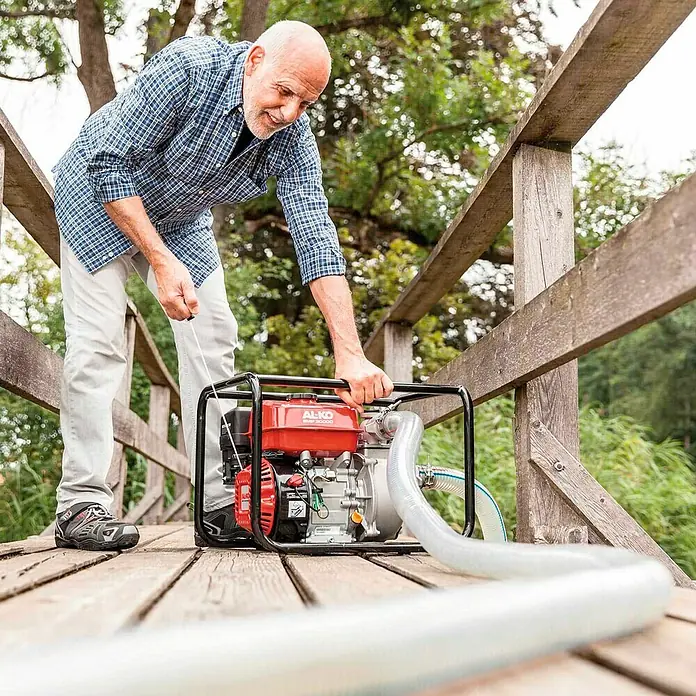 AL-KO Schmutzwasserpumpe - 4,1 kW, Max. Fördermenge: 30.000 l/h, Eintauchtiefe: 6 m - Ein Mann startet eine AL-KO Benzin-Wasserpumpe auf einer Holzbrücke. Die Pumpe ist rot und schwarz und hat einen Schlauch.