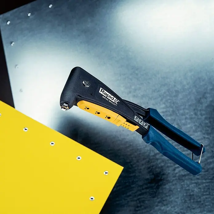 Rapid Remachadora RP40 MULTI - Apto para: Remaches de aluminio/acero inoxidable (Ø 3,2 mm/4 mm), Diámetro de remache: 4,8 mm, Mango ergonómico - Primer plano de una remachadora Rapid RP40 Multi con mango azul y rueda de ajuste amarilla frente a piezas de trabajo.