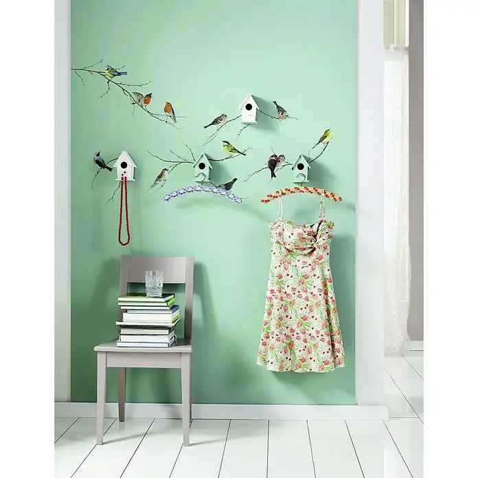 Komar Dekosticker Vogerl - Bunt, 50 x 70 cm - Helle und freundliche Raumecke mit einem floralen Kleid am Kleiderbügel, einem Stuhl mit Büchern und einem Glas, sowie einer Wanddekoration mit Vogelmotiven.