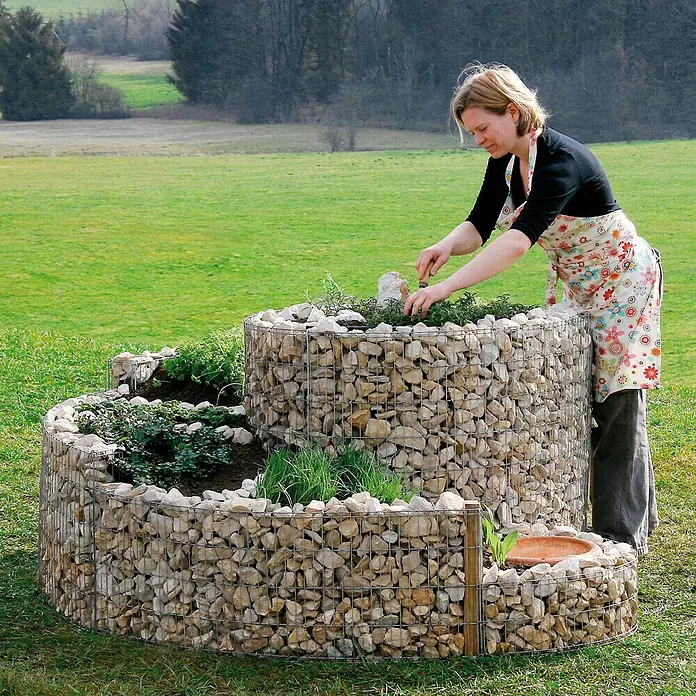 Bellissa Gabionen-Kräuterspirale - 150 x 200 x 80 cm, Stahl, Fassungsvermögen Erde: 1.350 kg - Eine Frau bepflanzt eine spiralförmige Kräuterschnecke aus Gabionen im Garten, ein attraktiver und praktischer Blickfang.