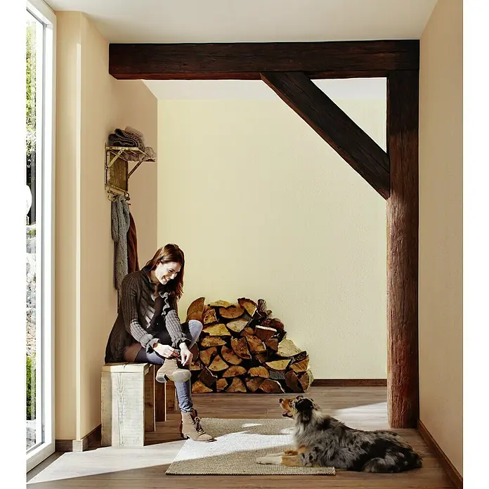 Knauf Putz-Abtönfarbe Compact Color - Terracotta, 6 g - Frau sitzt auf Bank und schnürt Stiefel in Diele mit Holzbalken, neben ihr ein Hund auf einem Teppich.