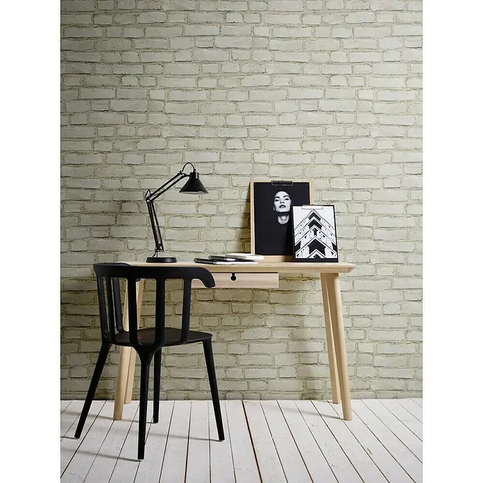 A.S. Création Neue Bude 2.0 ED II Vliestapete Steine - Natur, Steinoptik, 10,05 x 0,53 m - Schwarzer Stuhl und Schreibtisch mit Lampe, Notizbuch und zwei gerahmten Bildern vor einer Ziegelwand.