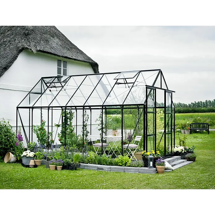 Halls Gewächshaus Magnum 148 - Außenmaß inkl. Dachüberstand (B x T): 257 x 446 cm, Grün, Wandstärke: 3 mm - Ein Gewächshaus mit schwarzem Rahmen und Glasplatten, umgeben von einem blühenden Garten mit Tisch und Stühlen.