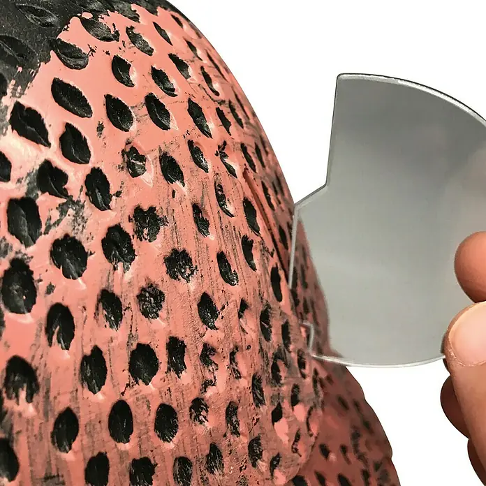 Altadex Búho Espanta pájaros - Altura: 41 cm - Primer plano de una mano que sostiene un calibrador curvo y reflectante contra un objeto rosa salpicado de negro. Énfasis en la precisión.
