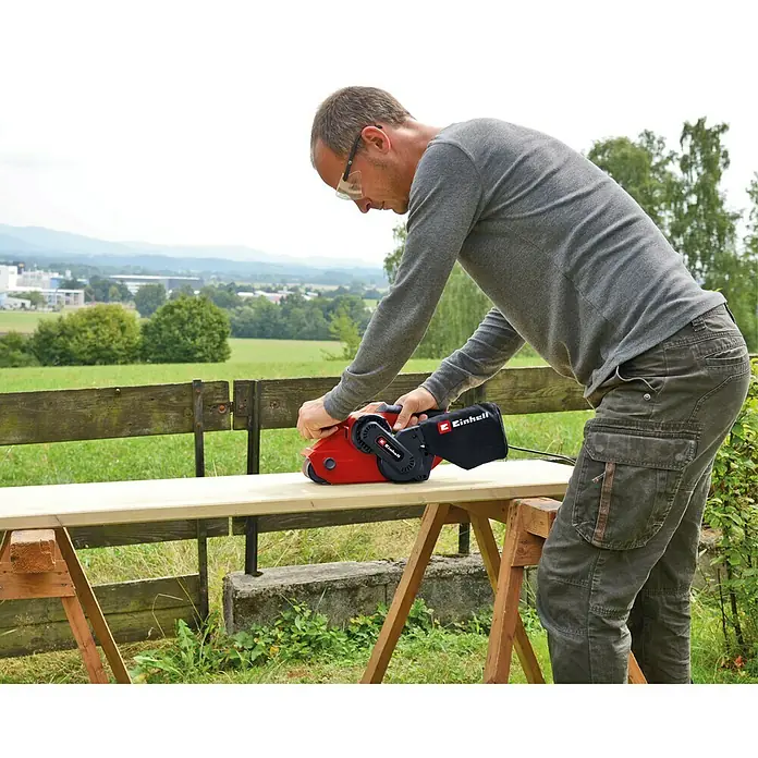 Einhell Elektro-Bandschleifmaschine TC-BS 8038 - 800 W - Ein Mann schleift ein Holzbrett im Freien mit einem roten Einhell Bandschleifer. Die Umgebung ist ländlich mit Bäumen und Bergen im Hintergrund.