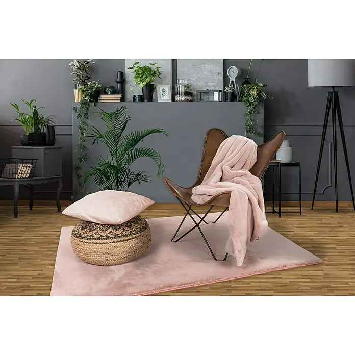 Decke Happy - Puderrosa, 200 x 150 cm, 100 % Polyester - Ein stilvolles Wohnzimmer mit einem flauschigen rosa Teppich, einem Ledersessel und vielen Zimmerpflanzen.