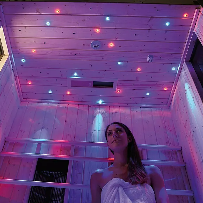Weka Farbvisionen Set B - Anzahl LED: 43 Stk., Mindestraumhöhe: 240 cm - Ein entspannender Moment in einer Infrarotsauna mit Farbtherapie und einer Frau, die in ein Handtuch gehüllt ist.