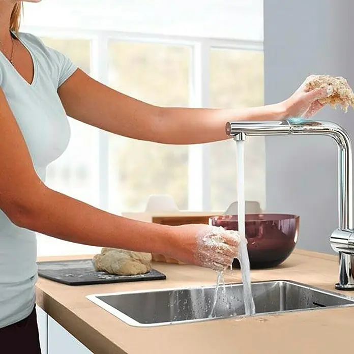 Grohe Spültischarmatur Minta Touch - Chrom, Glänzend - Eine Frau wäscht sich die Hände an einem modernen, chromfarbenen Wasserhahn in einer hellen Küche mit Holzoberflächen.