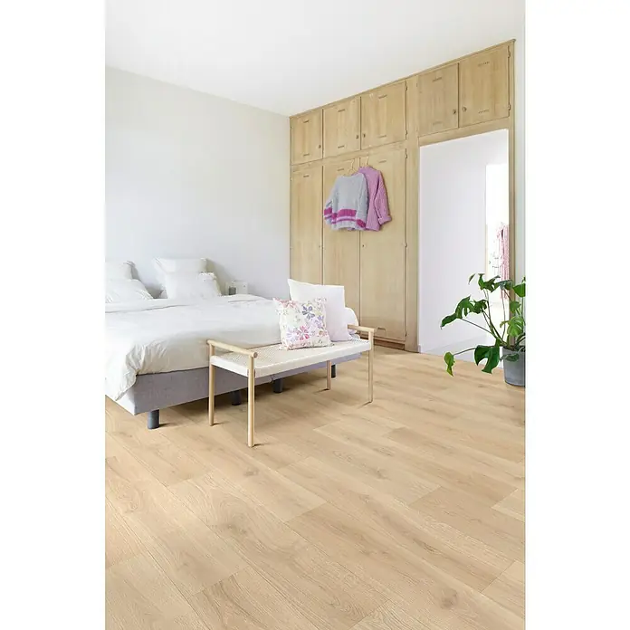 Beauflor PVC-Boden Meterware Jupiter - 400 cm, Beige, Wood 20 - Einladendes Schlafzimmer mit hellem Holzboden, Bett mit vielen Kissen und einer Bank davor. Ein großer Schrank und eine Zimmerpflanze runden das Bild ab.