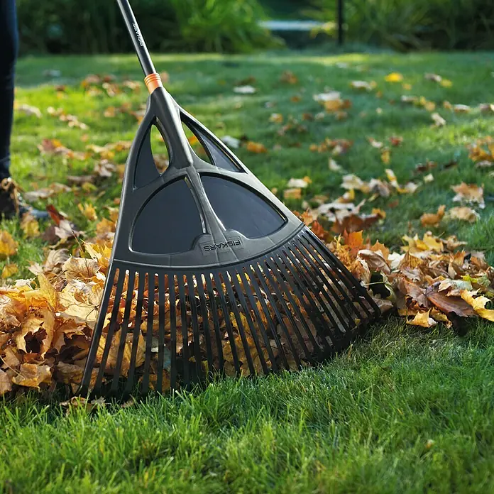 Fiskars Solid Laubbesen XL - Arbeitsbreite: 65 cm, Anzahl Zinken: 27, Langstiel - Eine schwarze Fiskars Laub Harke liegt auf einem Rasen voller Herbstlaub und ist bereit zum Aufräumen. Perfekt für Gartenarbeiten.