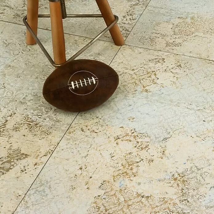 Feinsteinzeugfliese Carpet Natural - 50 x 100 cm, Sand, Matt - Brauner American Football mit weißen Schnürsenkeln auf einem gemusterten Fliesenboden neben hölzernen Stuhlbeinen.