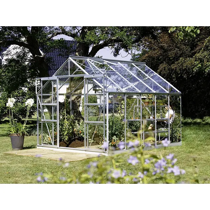 Vitavia Gewächshaus Mars 8300 - Außenmaß inkl. Dachüberstand (B x T): 257 x 321 cm, 3 mm, Aluminium - Ein Gewächshaus aus Glas und Metall steht im Garten, umgeben von Grünpflanzen. Eine Frau ist darin zu sehen, die sich um die Pflanzen kümmert.