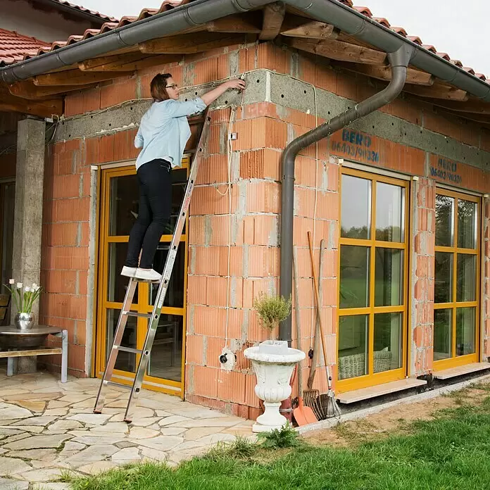 Krause Monto Escalera de apoyo - Número de peldaños: 9 ud., Altura de trabajo: 3,6 m, Carga soportada: 150 kg, Aluminio - Una mujer en una escalera multiusos plateada instala cables en la pared exterior de una casa en construcción.