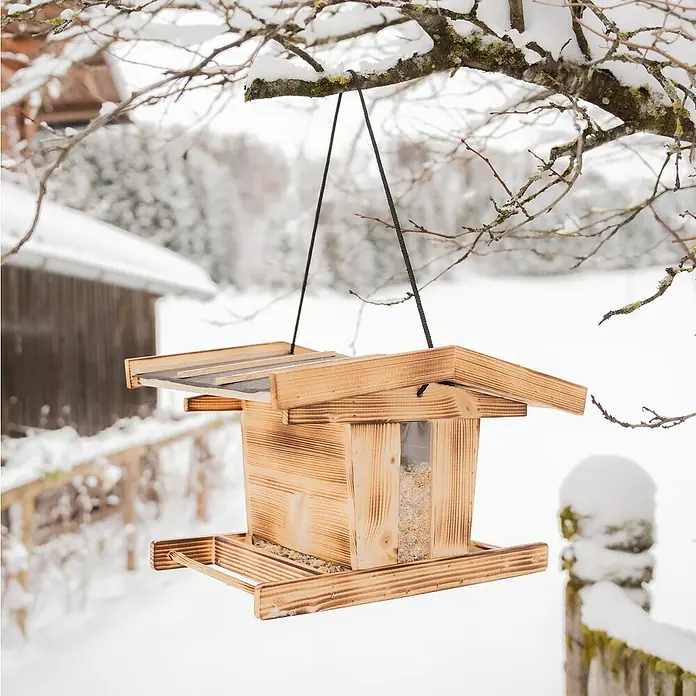Windhager Vogelfutterhaus Canteen XL - L x B x H: 29 cm x 38 cm x 23,5 mm, Braun, Kiefer - Ein hölzernes Vogelhaus hängt an einem verschneiten Ast, gefüllt mit Futter, ideal für Wintervögel.
