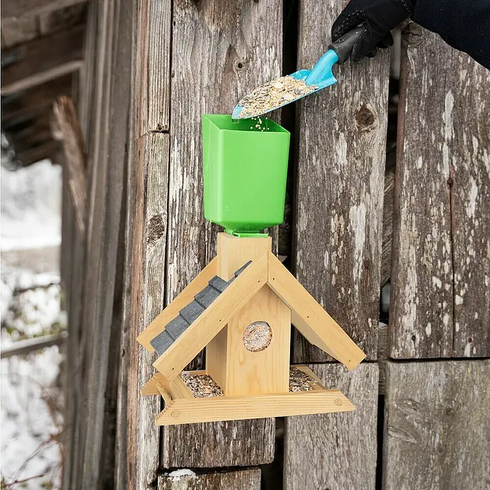 Windhager Vogelfutterhaus Finca - 16,5 x 29 x 25 cm, Braun, Holz, Kiefer - Eine Person füllt einen Futterspender für Vögel mit einer blauen Schaufel auf, der an einer Holzwand befestigt ist.