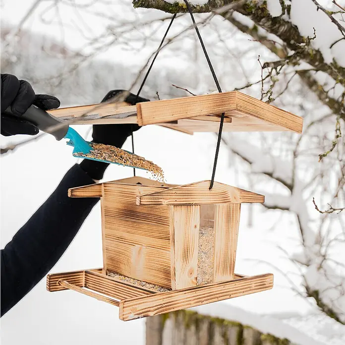 Windhager Vogelfutterhaus Canteen XL - L x B x H: 29 cm x 38 cm x 23,5 mm, Braun, Kiefer - Eine Person befüllt einen hölzernen Vogelhäuschen mit einem blauen Schaufel vor einem verschneiten Hintergrund.